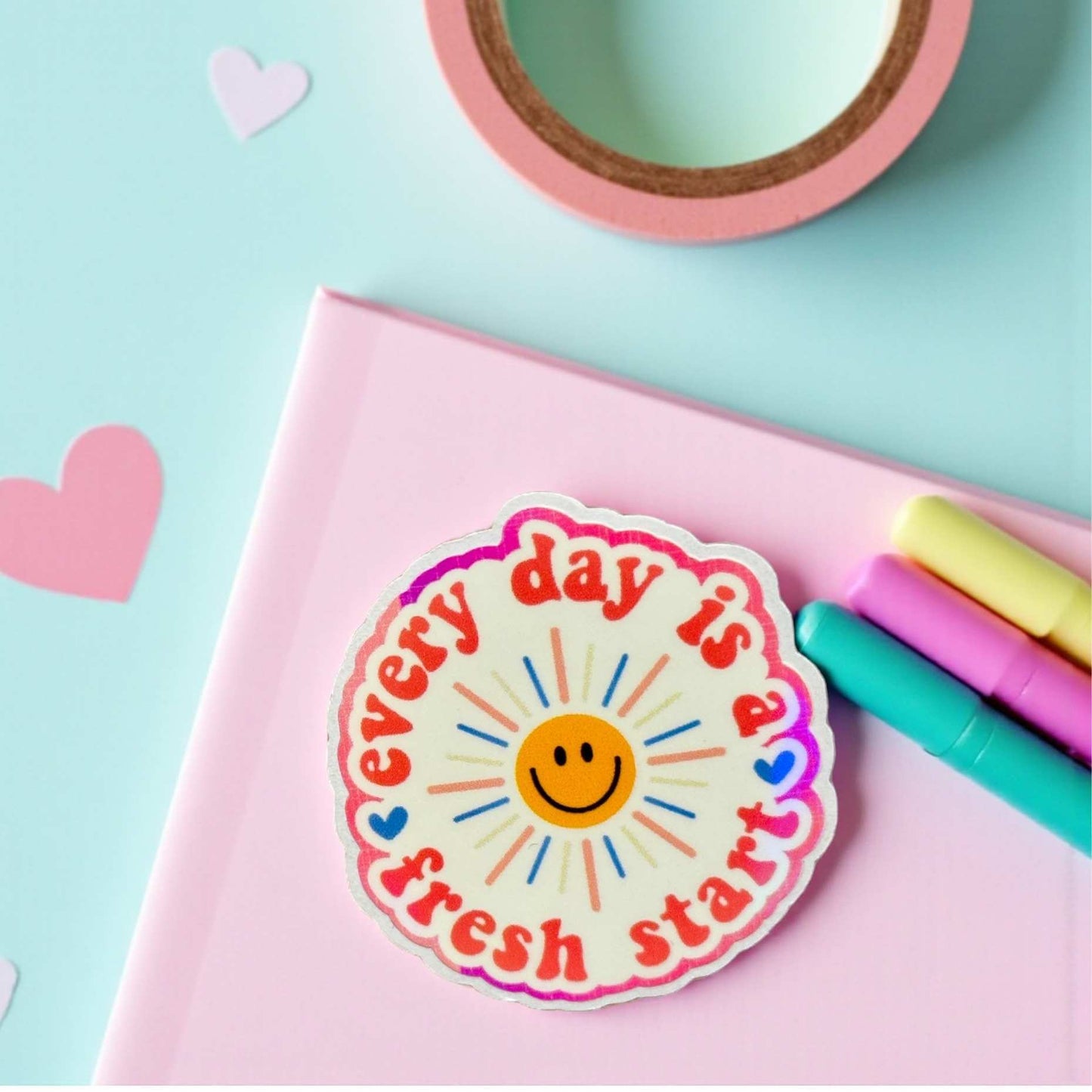 Colorful sticker with 'Every day is a fresh start' text on a pink notebook with stationery items.