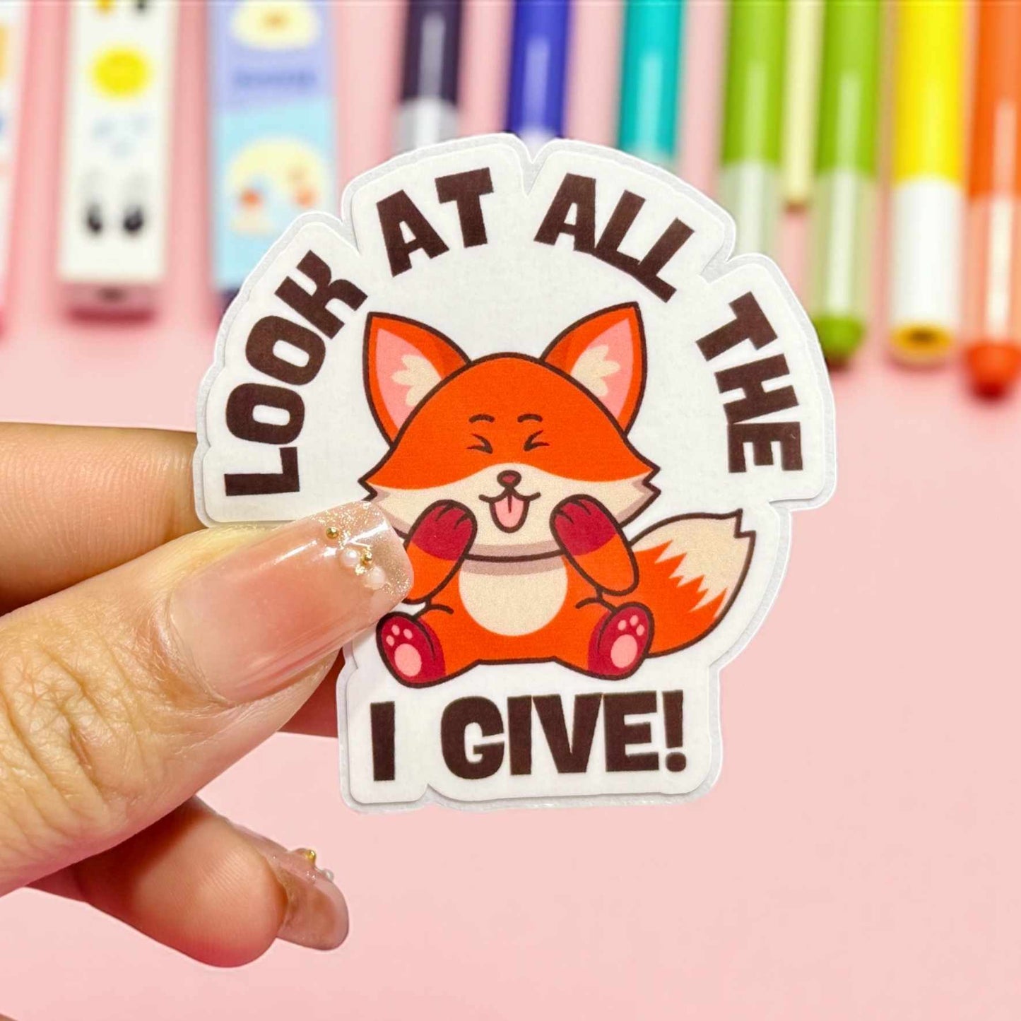 Sticker with a fox design and text held by a hand against a colourful background
It says look at all the FOX PHOTO I give