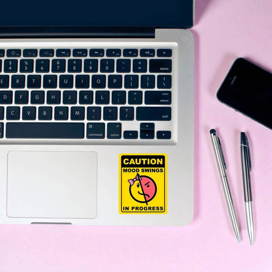 Laptop with a 'Caution: Mood Swings in Progress' sticker on a pink surface with pens and a phone.