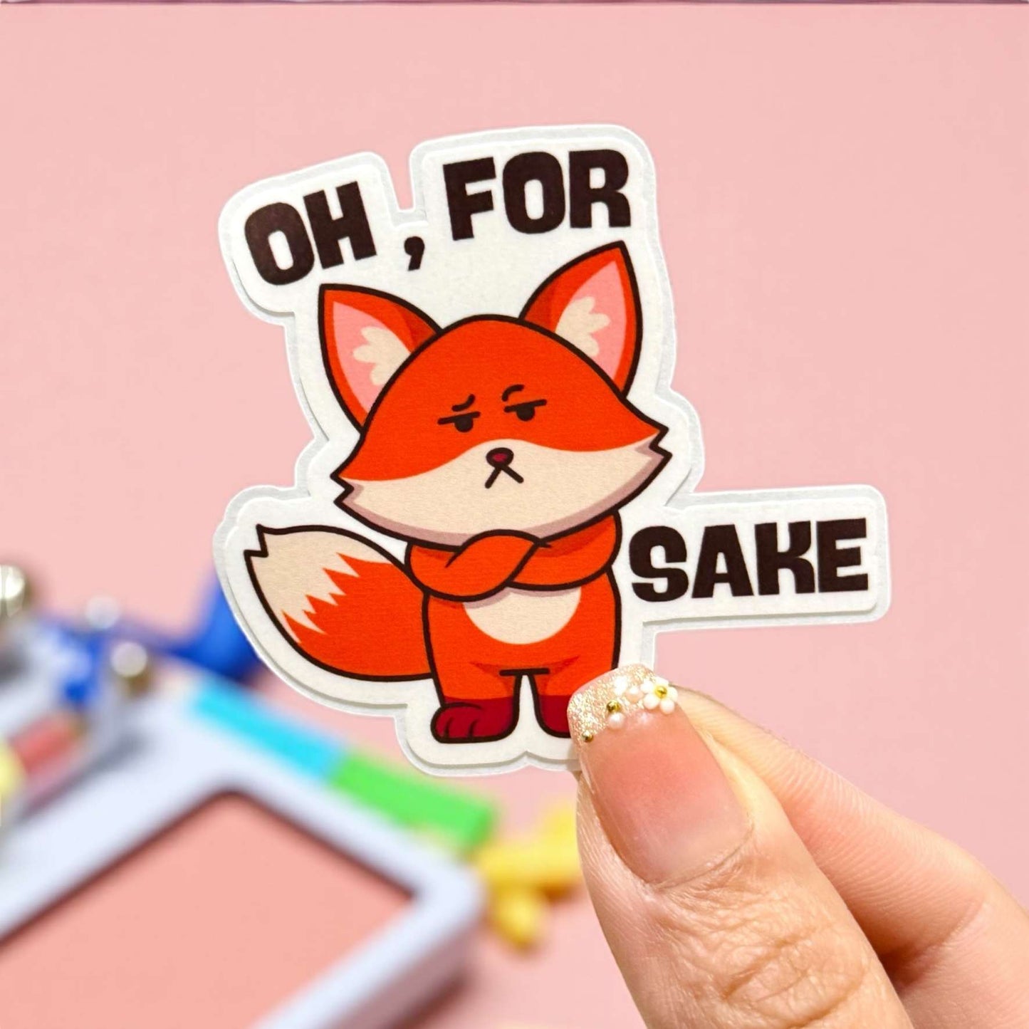 Sticker with a fox and text 'OH, FOR SAKE' held by a hand on a pink background