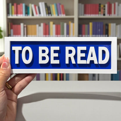 Hand holding a sign that says 'TO BE READ' in front of a bookshelf.