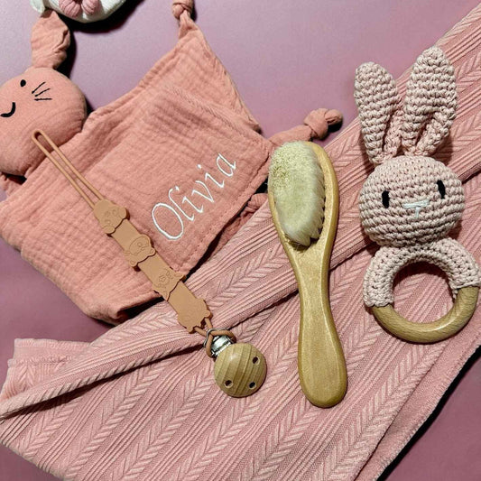 Baby care items including a pacifier clip, brush, and rattle on a pink textured blanket with name embroidered.
