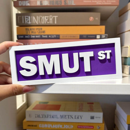Hand holding a sign with 'SMUT ST' in front of a bookshelf.
