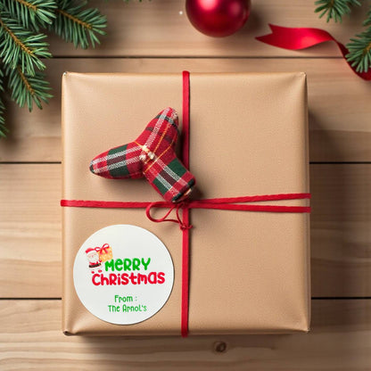 Gift wrapped in brown paper with a red ribbon and 'Merry Christmas' label with customisation on a wooden surface with Christmas decorations.