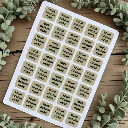 Sheet of customizable stickers with 'Your Design Here' text on a wooden surface with greenery.