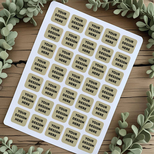 Sheet of customizable stickers with 'Your Design Here' text on a wooden surface with greenery.