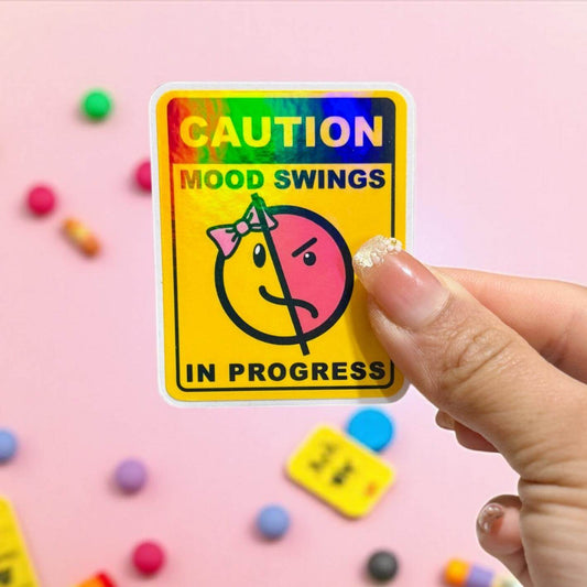 Hand holding a colorful sticker with 'Caution: Mood Swings In Progress' text on a pink background with small colorful objects.