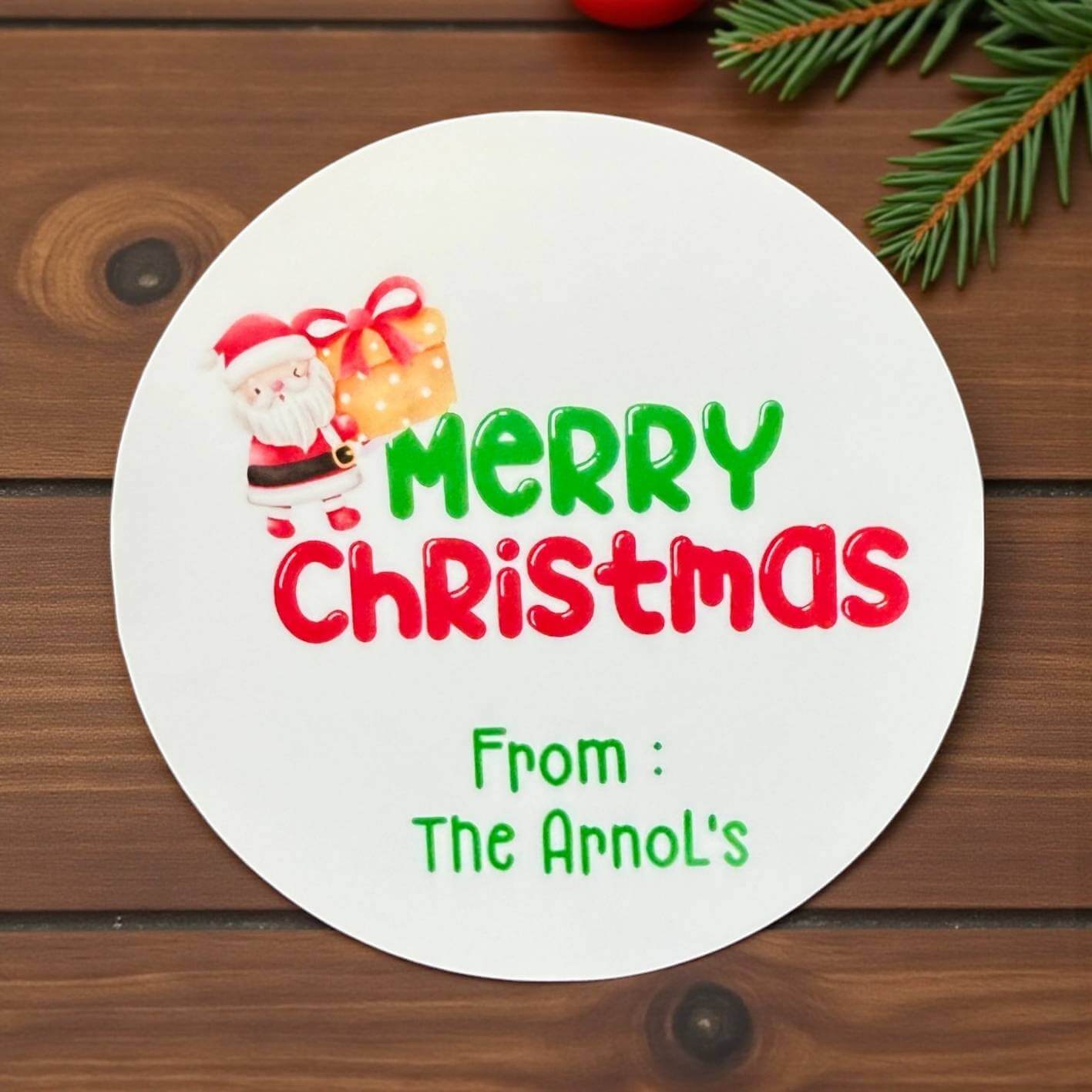 Round white card with Santa Claus and gift, 'Merry Christmas' text, and personalised name on a wooden background.