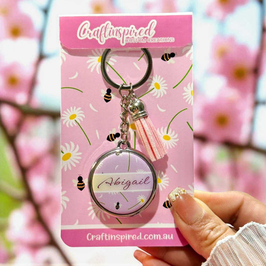 Keychain with custom name 'Abigail' on a pink packaging with floral design, held against a blurred floral background.