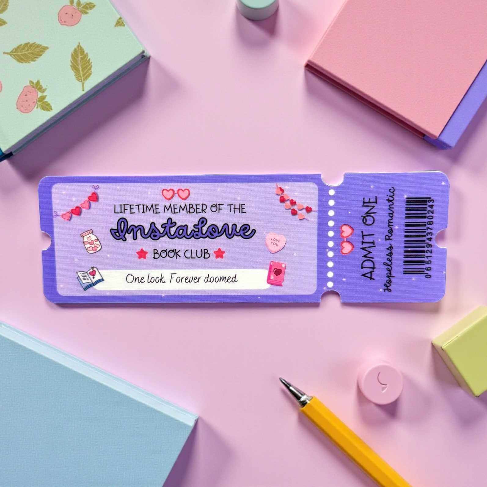 Purple book club membership bookmark ticket with decorative elements on a pink surface with stationery items.