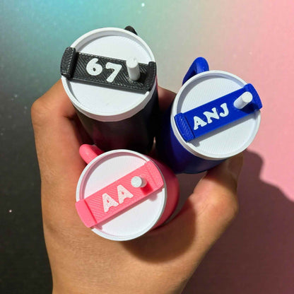 Three small cylindrical tumbler container keychains with colourful lids held in a hand against a gradient background.