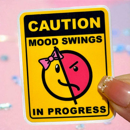 Sticker with 'Caution: Mood Swings in Progress' text and cartoon face on a pink background