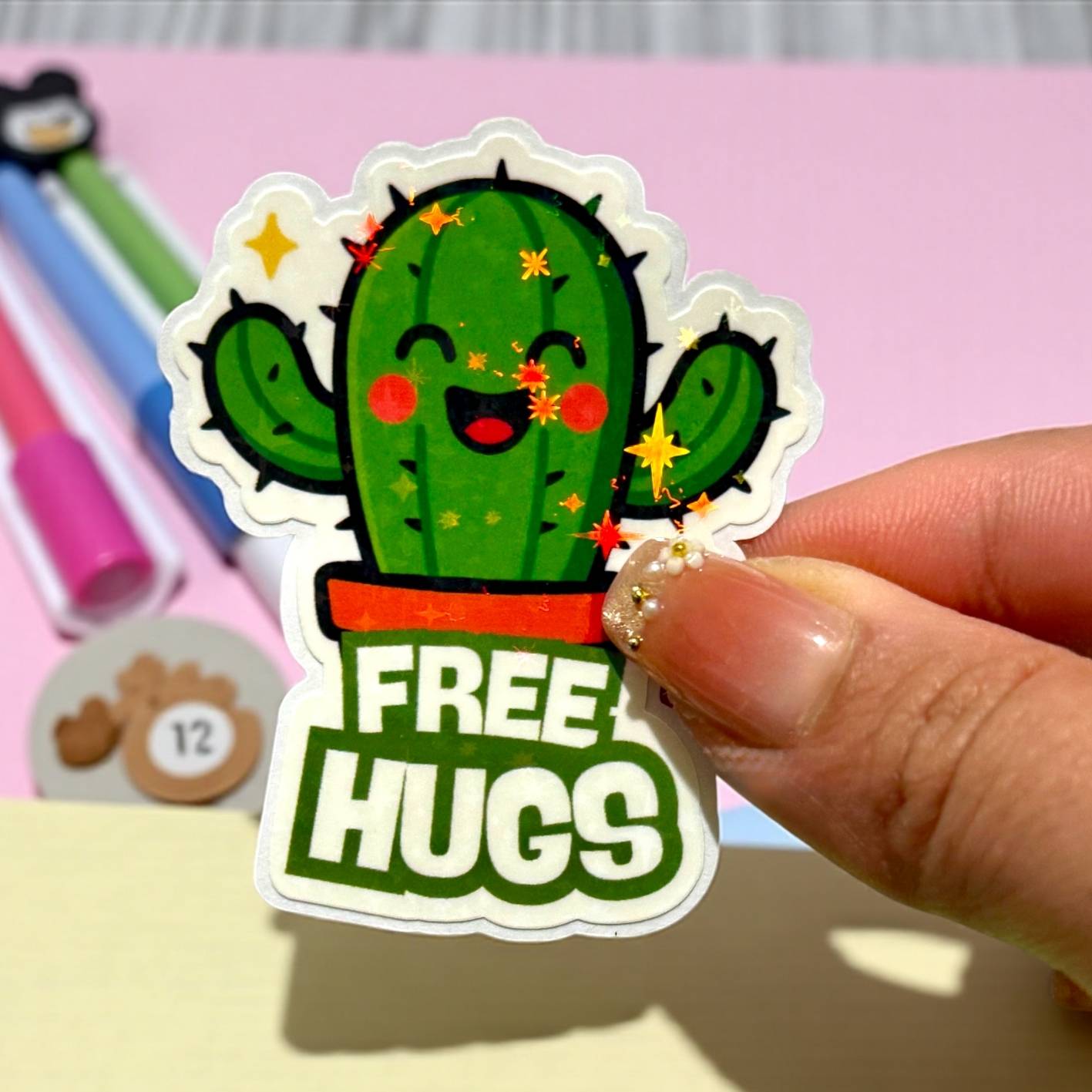 Sticker of a cactus with 'FREE HUGS' text held by a hand on a colorful background.