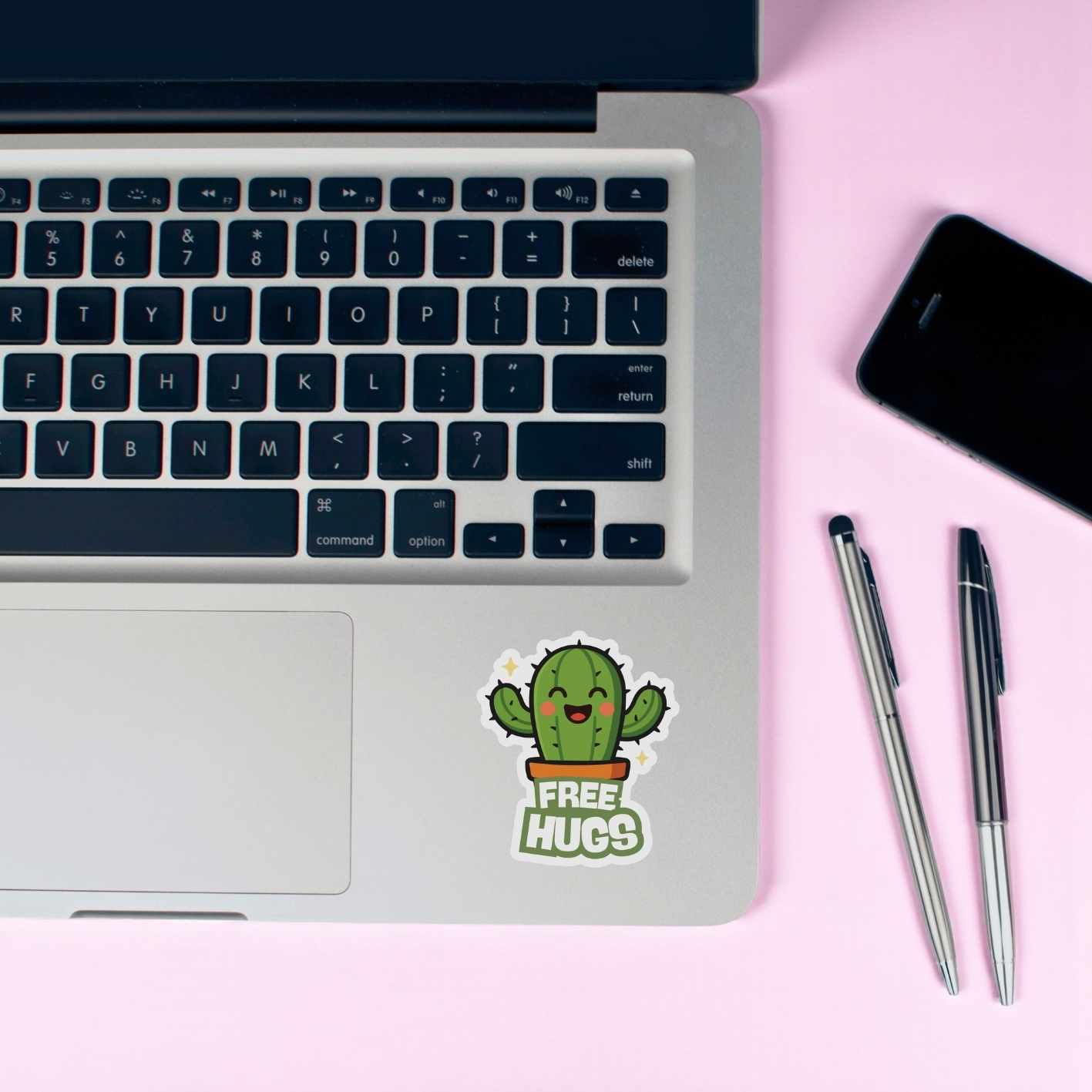 Laptop with a cactus sticker reading 'Free Hugs' on a pink surface with a phone and pens.